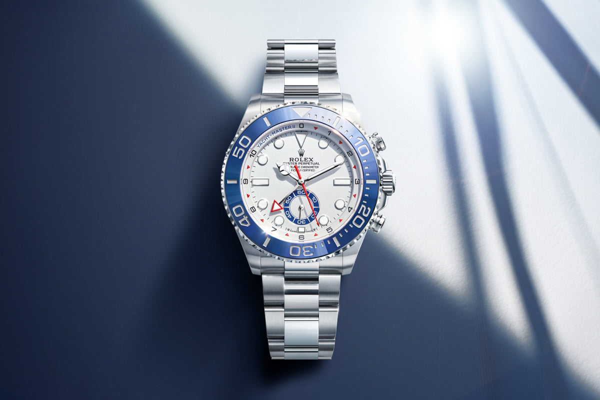Rolex Yacht-Master II watch with a steel bracelet, blue bezel and white dial on a reflective background.