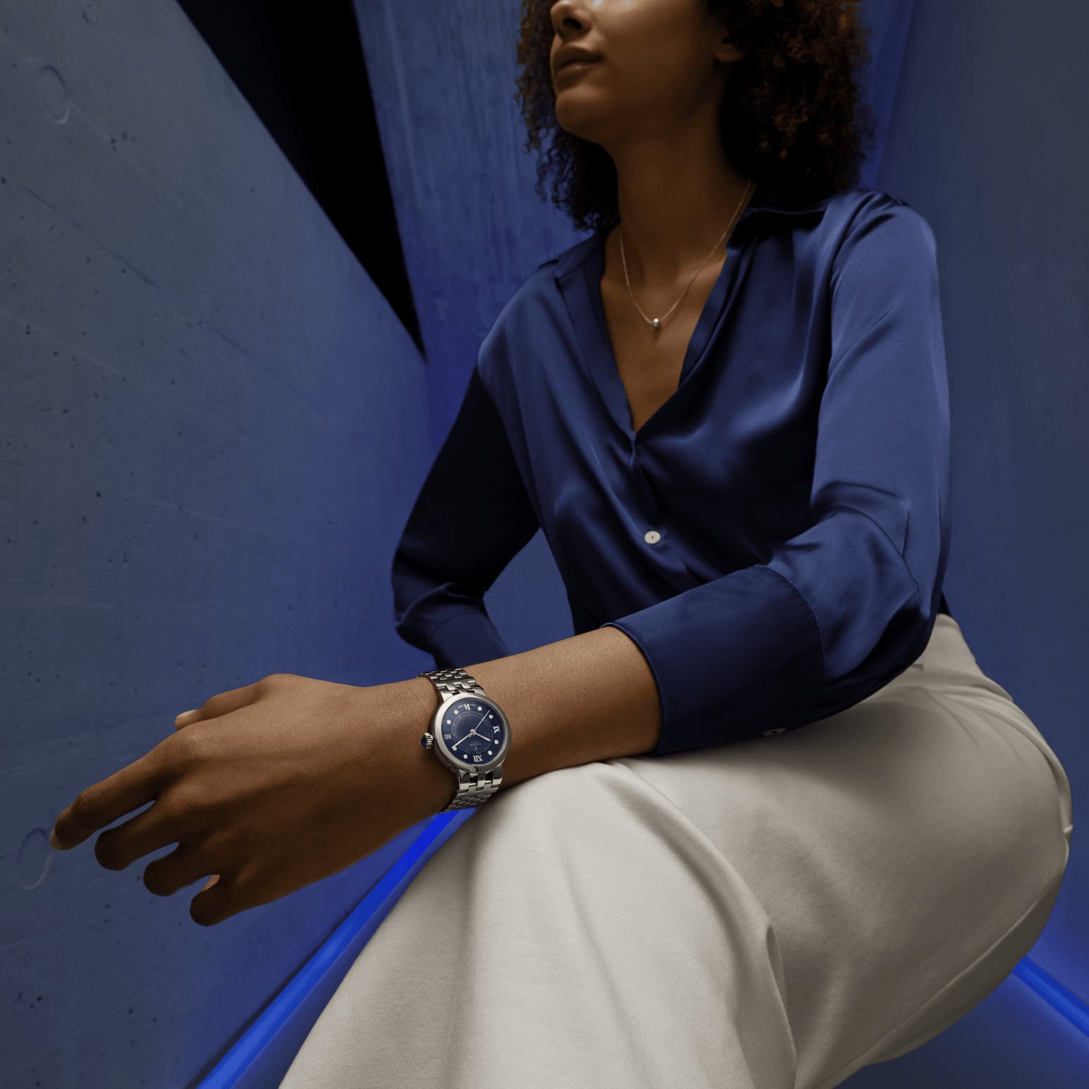 Woman wearing a blue shirt and beige trousers with a Tudor Clair de Rose watch, sitting against a blue wall.