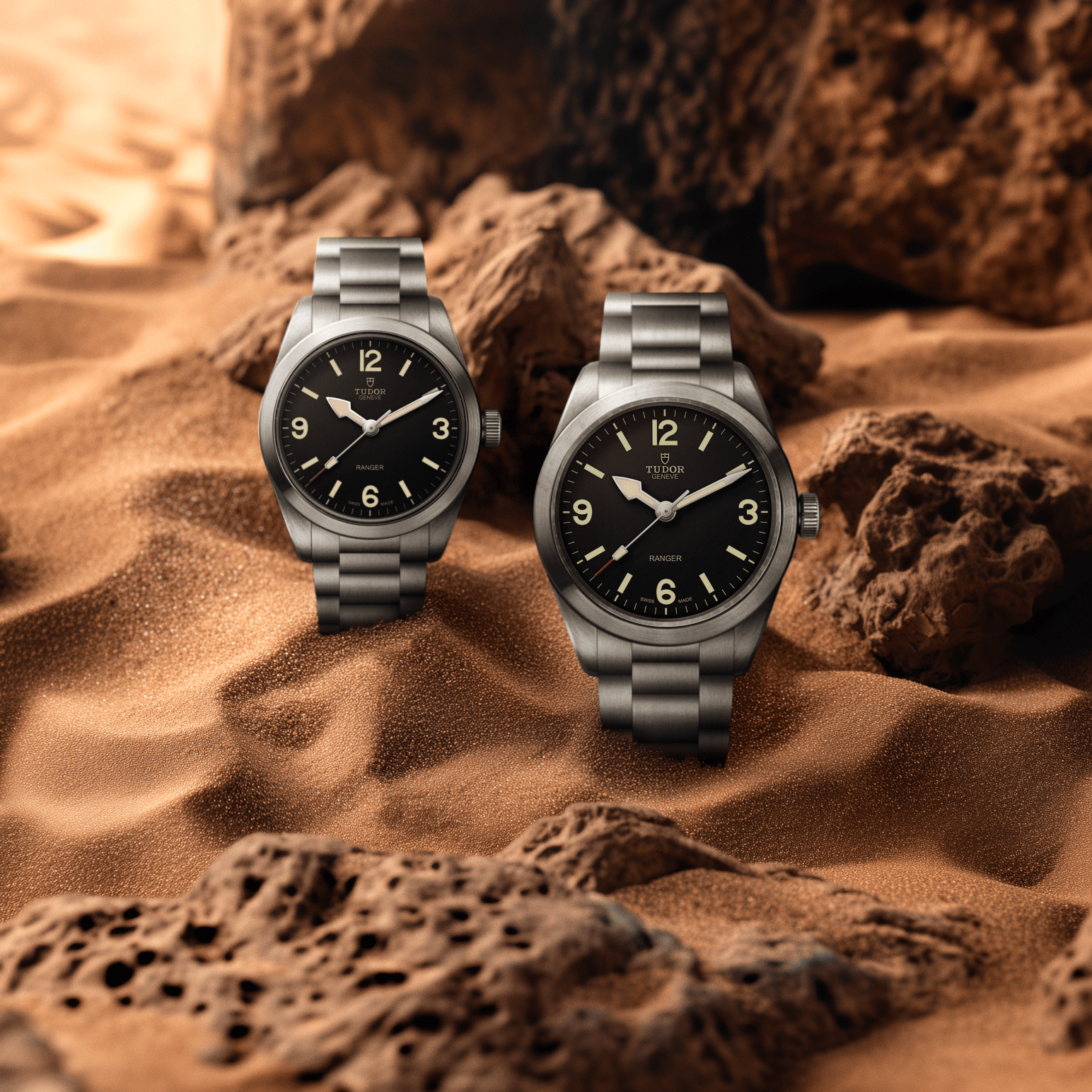Two Tudor Ranger watches with steel bracelets and black dials on a sand background.