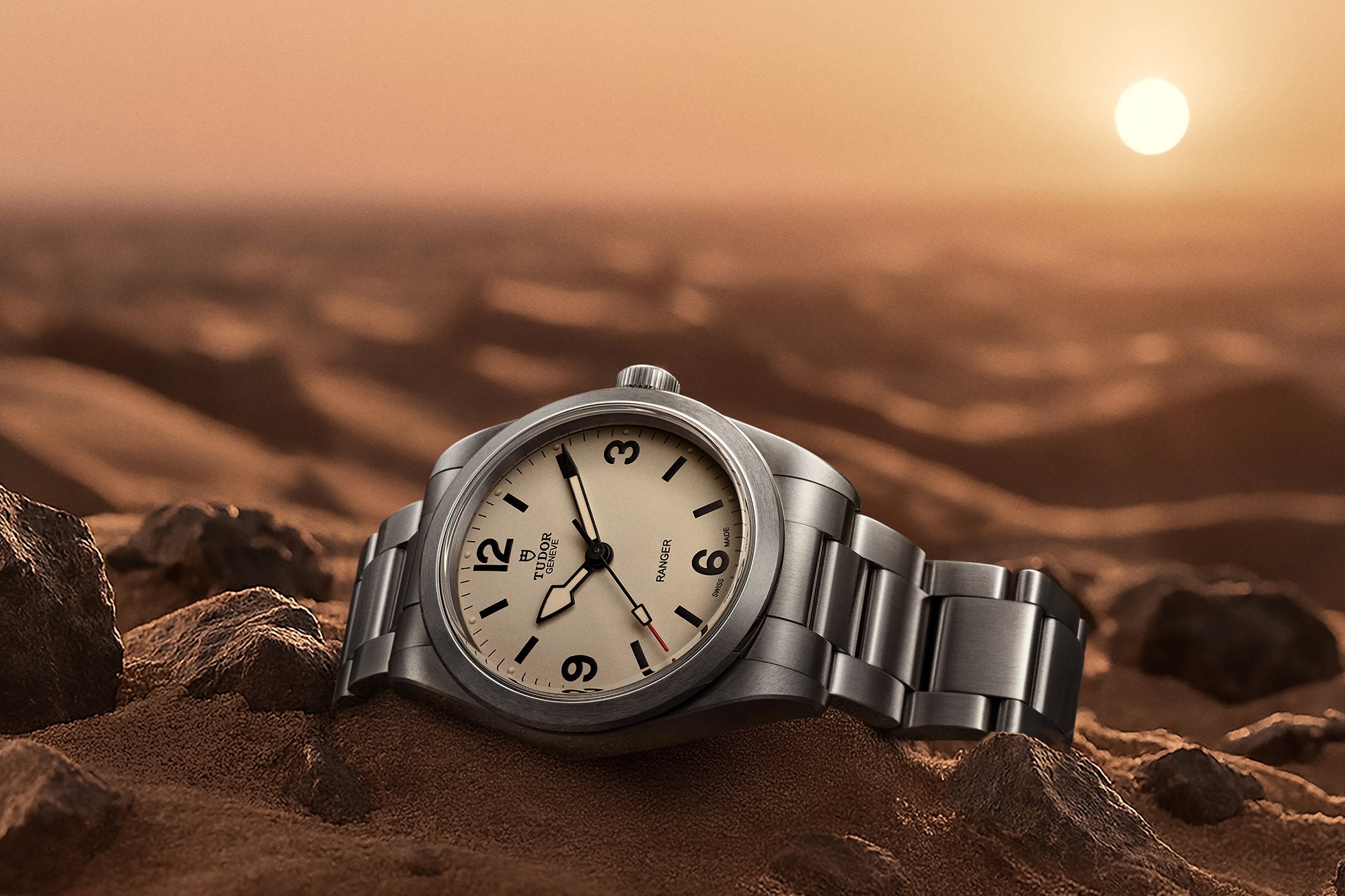 Tudor Ranger watch with a beige dial and steel bracelet, displayed on its side in the sand.