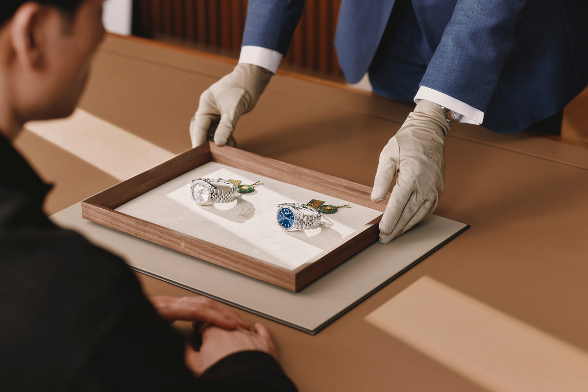 Person in a blue suit and white gloves presenting two watches on a wooden tray to another person.