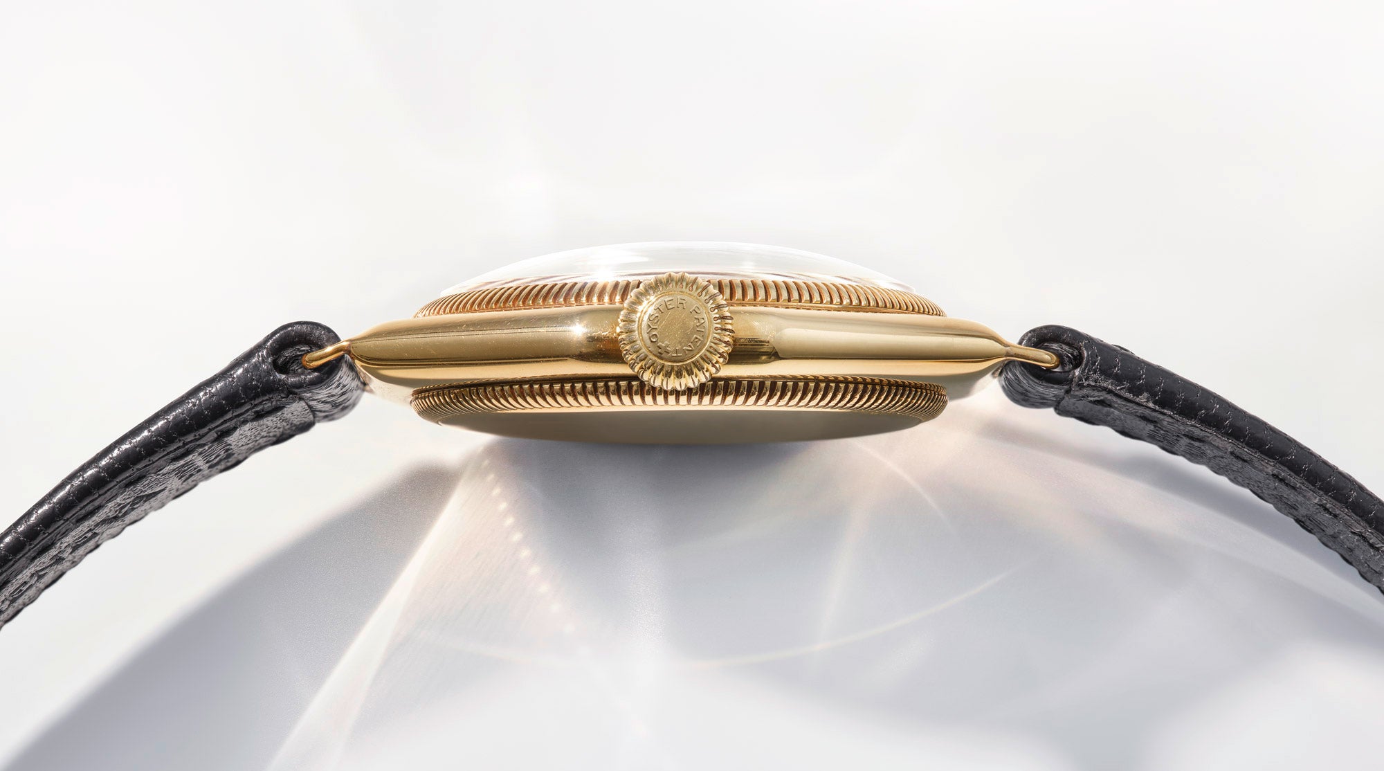 Side-profile of Rolex Oyster gold case with black leather strap on a reflective background.