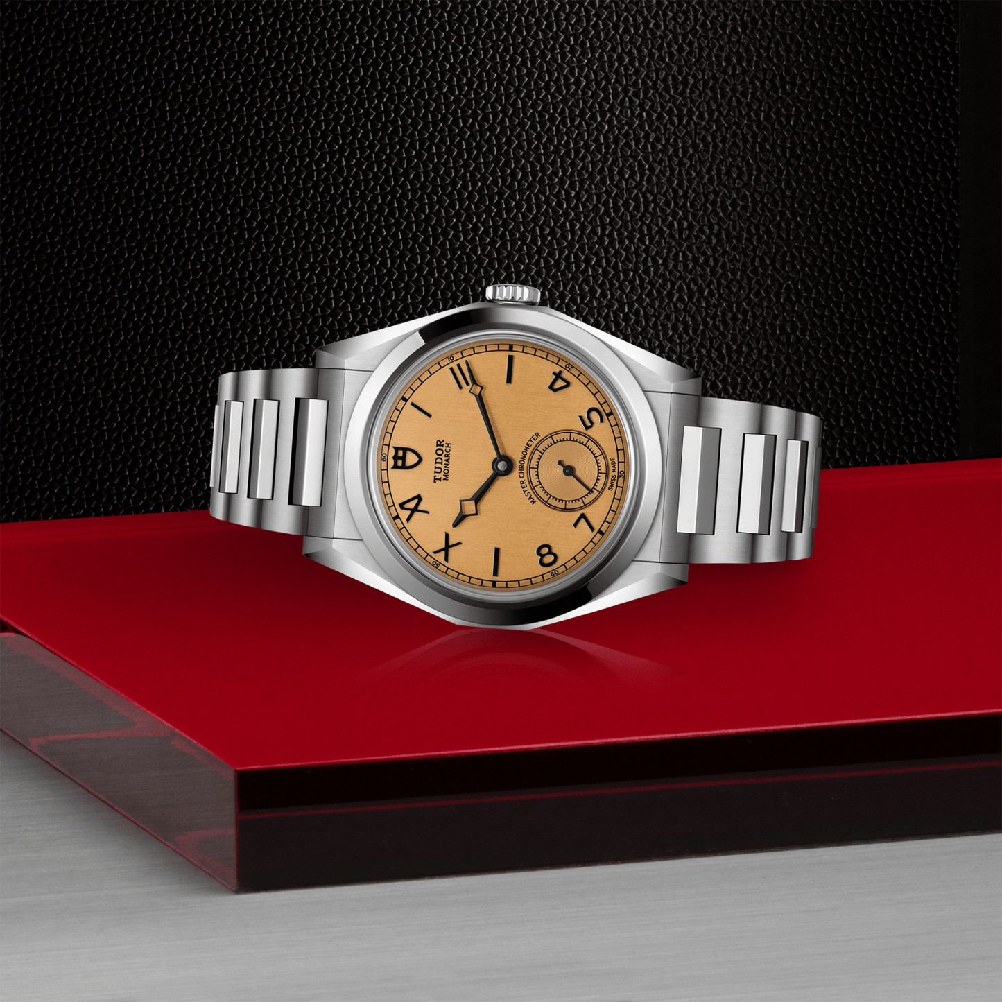 Tudor Monarch watch with steel bracelet and champagne dial on red platform against a black background.