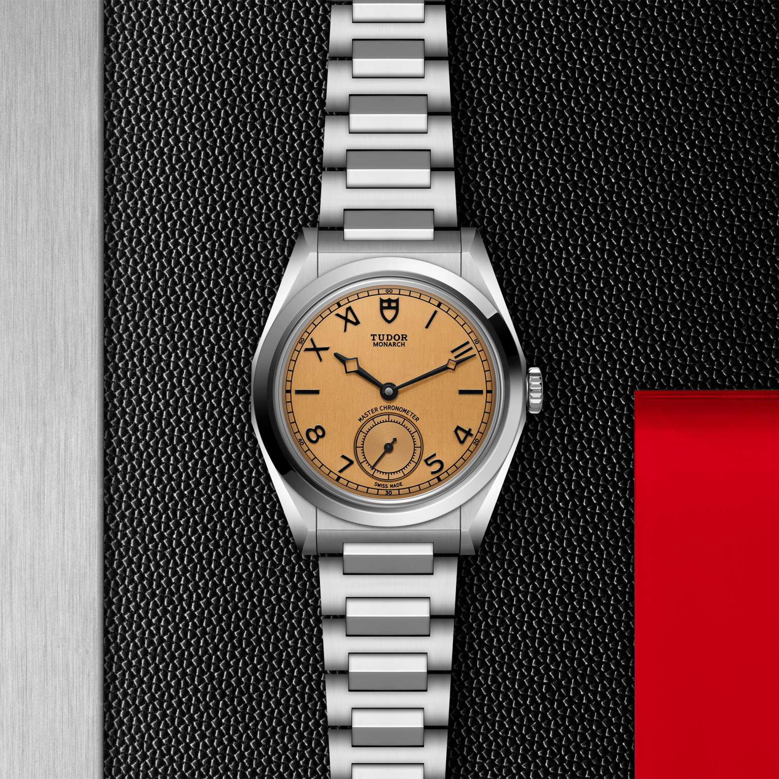 Tudor Monarch watch with steel bracelet and champagne dial on a black textured background.