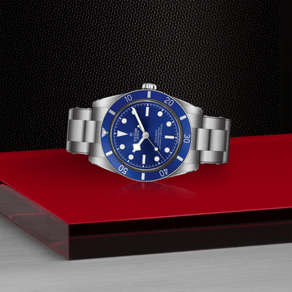 Tudor Black Bay 54 watch with a steel bracelet and blue dial on red platform against a black background.