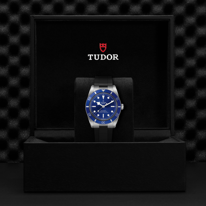 Tudor Black Bay 54 watch with a black rubber strap and blue dial in a black box against a black background.